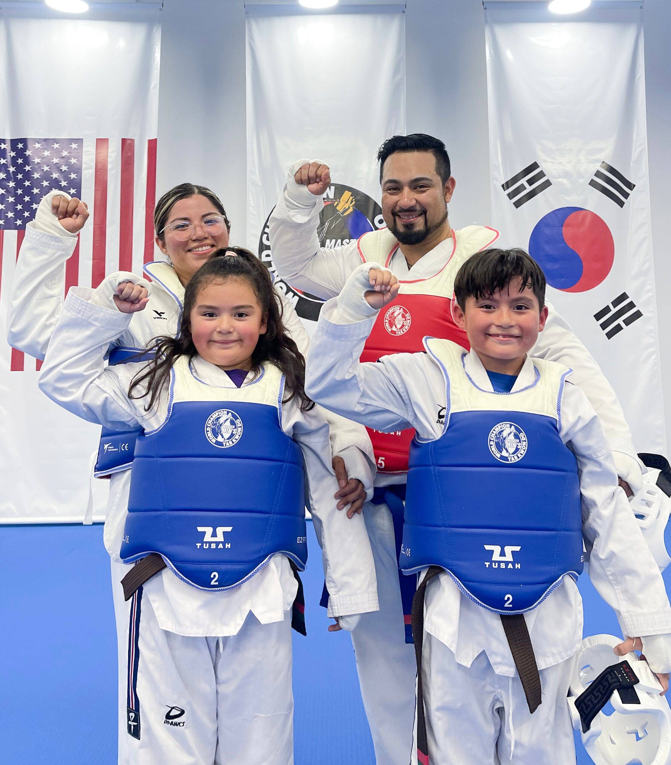 A parent and two children in sparring gear raising their fists in front of the flags
